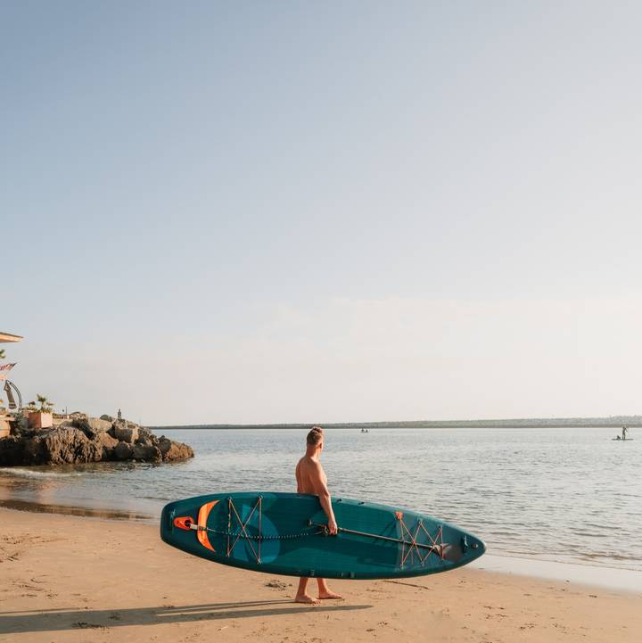 A person walks barefoot on a sandy beach, carrying a blue Weekender Tour Inflatable Stand Up Paddle Board, with calm waters and a jetty in the background.