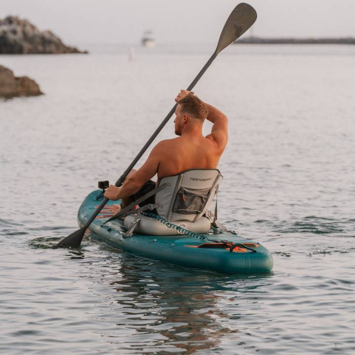 A shirtless man paddles the Weekender Tour Inflatable Kayak with an Inflatable Seat in calm water, with a distant boat and rocky shoreline visible under a serene sky.