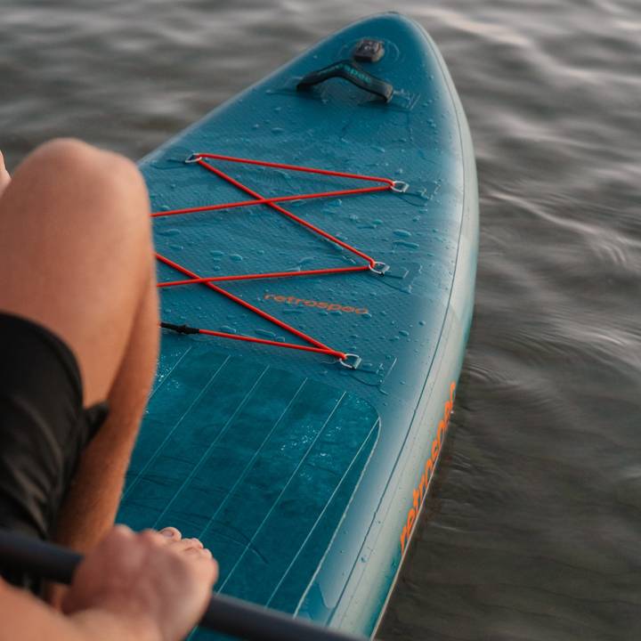 Close-up of a paddleboard with orange bungee cords, person's hand gripping the paddle, and water droplets on a blue surface.