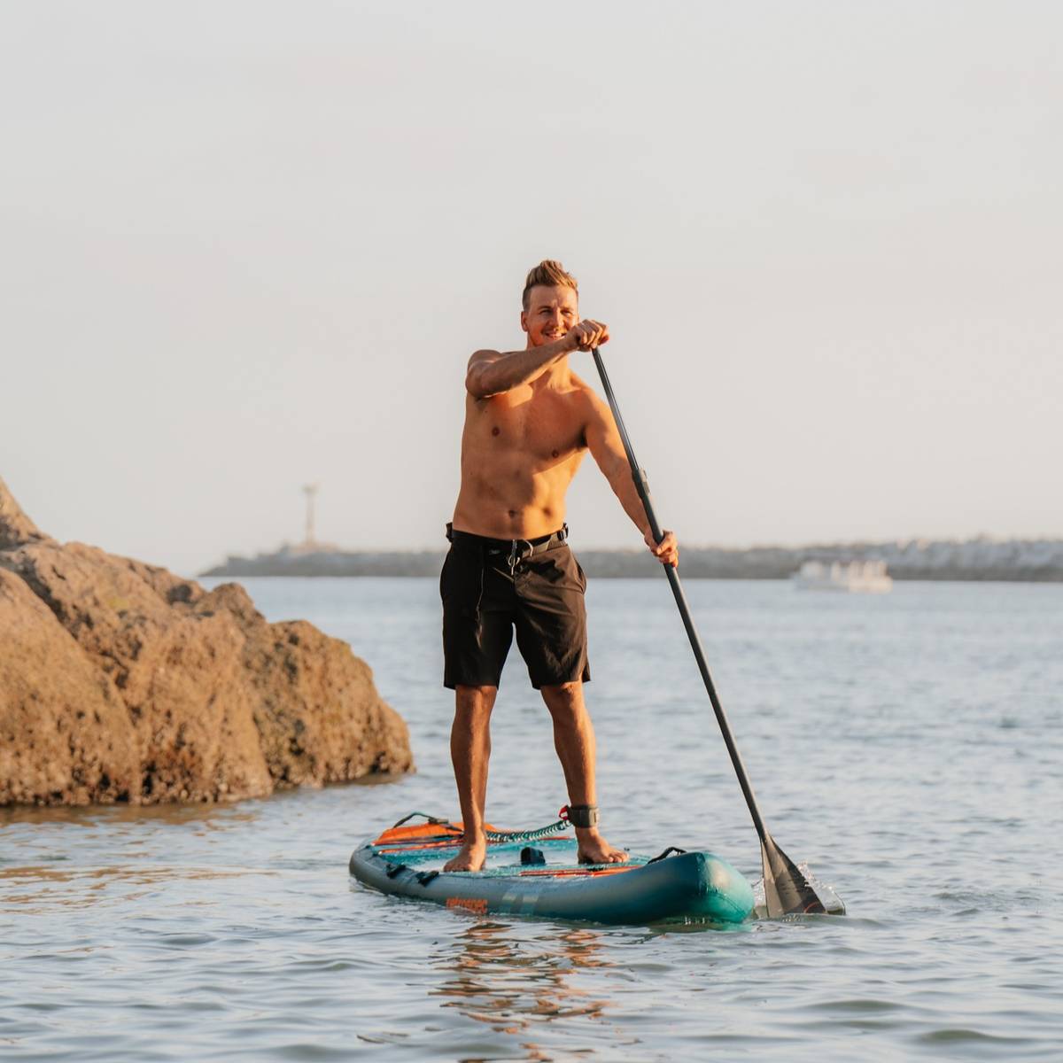 A man paddles on a stand-up paddleboard in calm waters, surrounded by rocks and a distant shoreline under a clear sky.