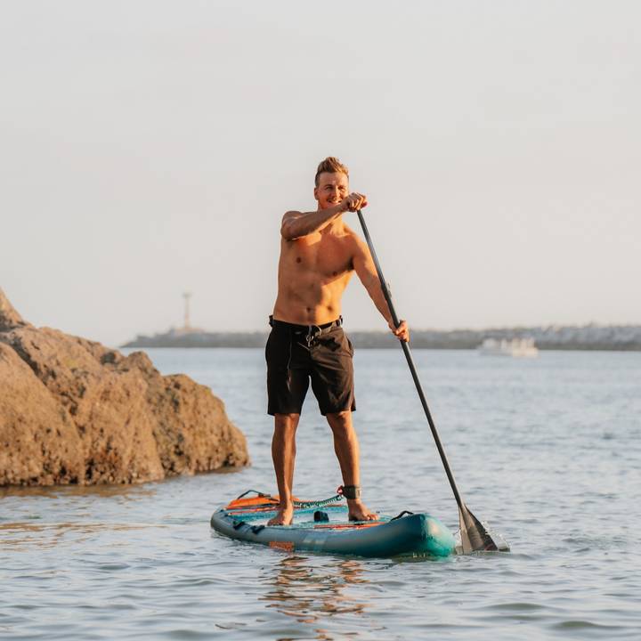 A man paddles on a stand-up paddleboard in calm waters, surrounded by rocks and a distant shoreline under a clear sky.