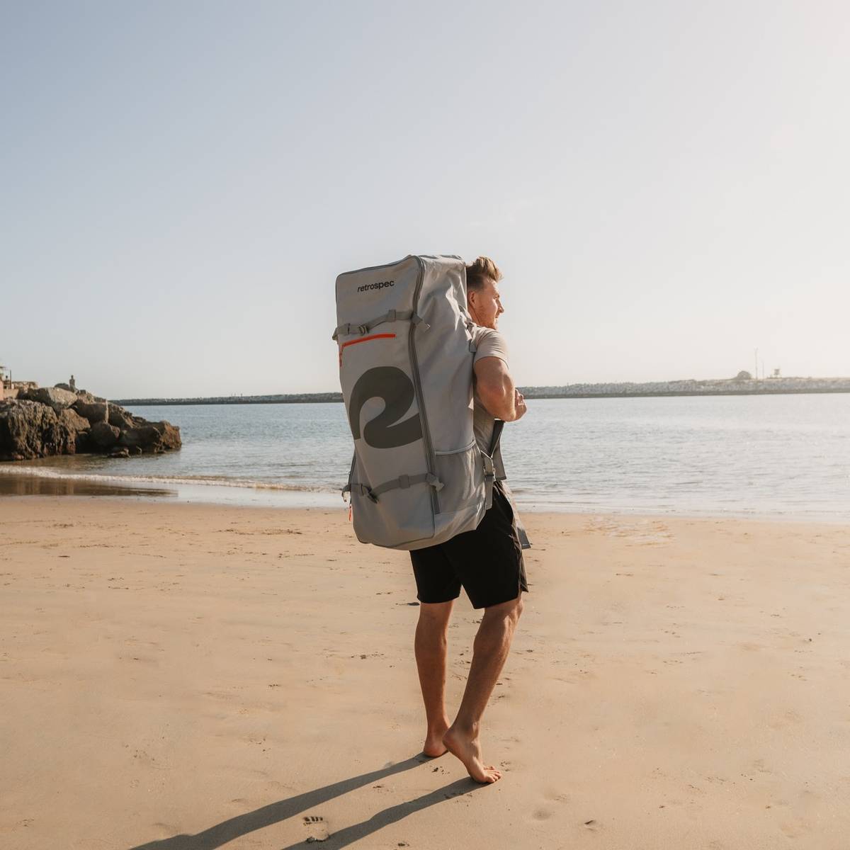 A person walks barefoot on a sandy beach, carrying a large gray backpack, with calm waters and a jetty in the background.