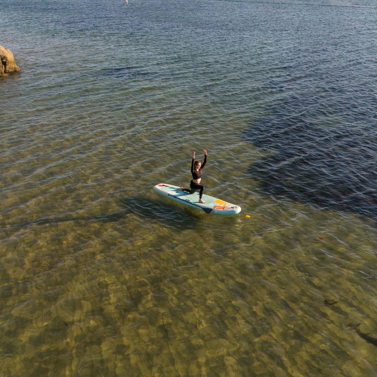 A person balances on a Weekender Yogi 10'8" Inflatable Stand Up Paddle Board with arms raised, gliding over clear, shallow water.