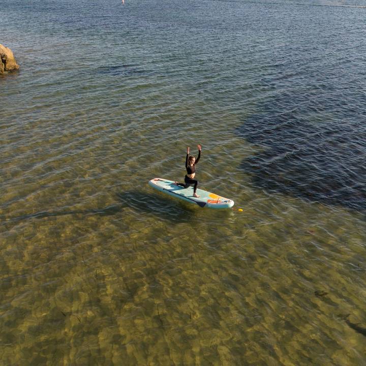 A person balances on a Weekender Yogi 10'8" Inflatable Stand Up Paddle Board with arms raised, gliding over clear, shallow water.