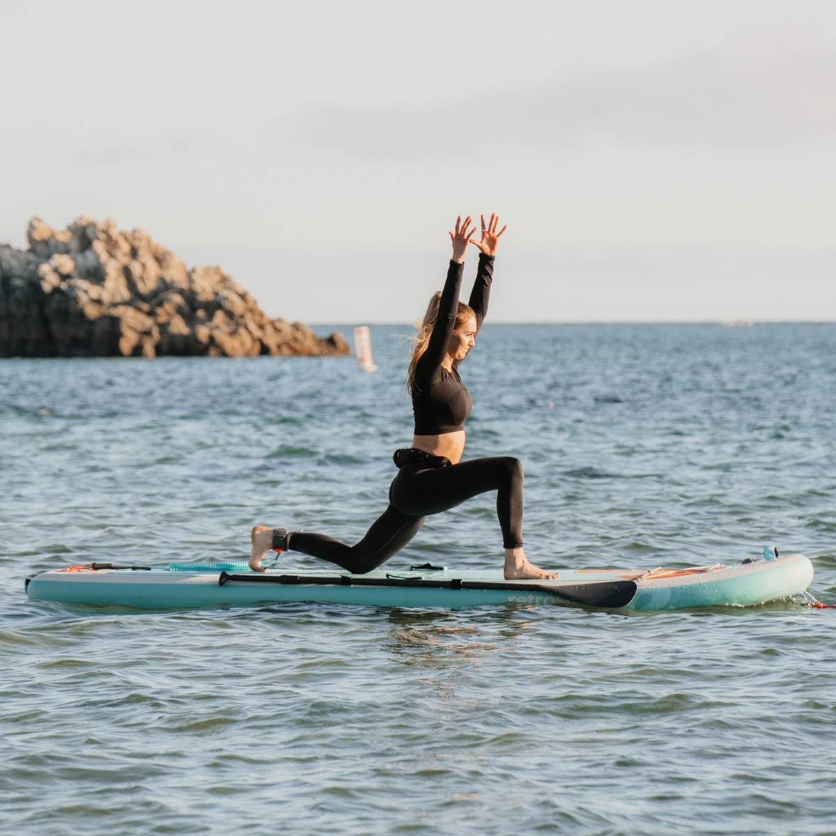 A person performs a yoga pose on a Weekender Yogi 10'8" Inflatable Stand Up Paddle Board in calm waters, with rocky formations in the background under a clear sky.