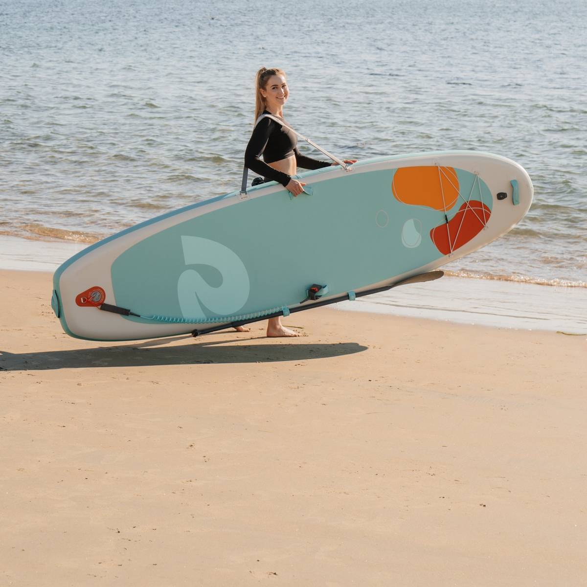 A person stands on a sandy beach, carrying a colorful Weekender Yogi 10'8" Inflatable Stand Up Paddle Board, with calm waters in the background and a clear blue sky above.