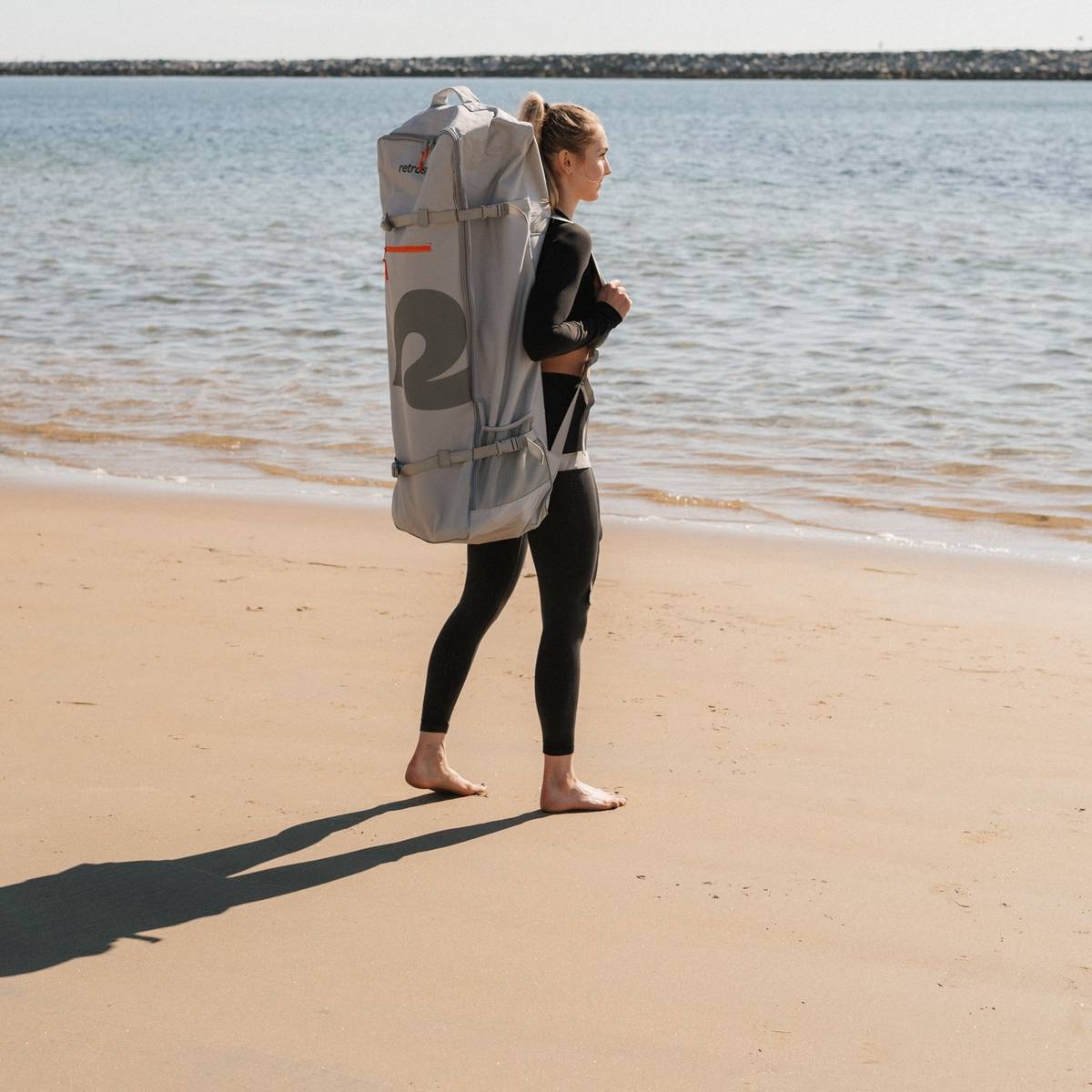 A person wearing a black outfit carries a large, gray backpack along a sandy beach, with calm water in the background reflecting sunlight.
