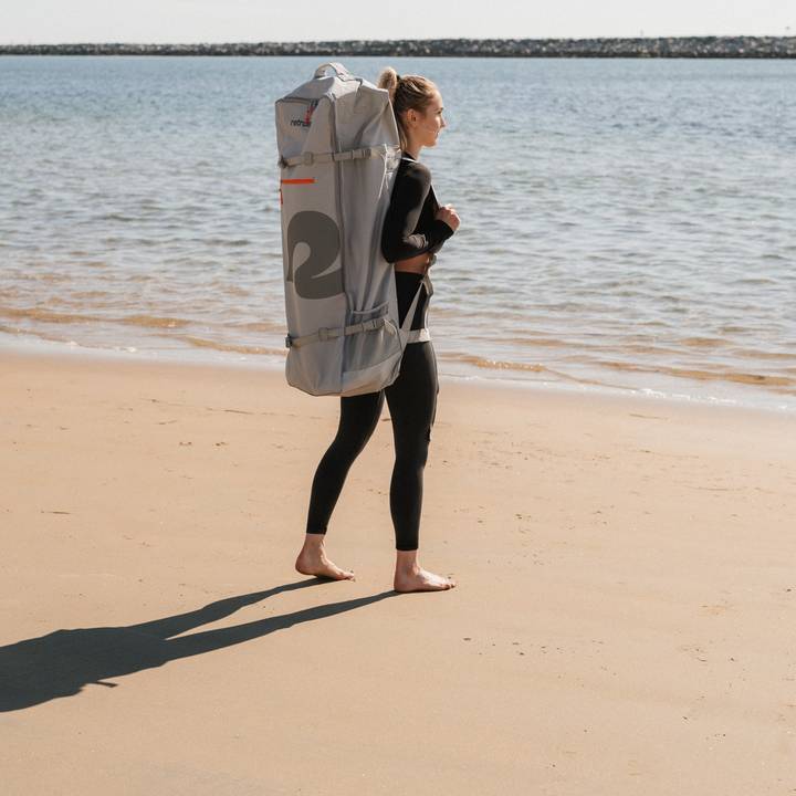 A person wearing a black outfit carries a large, gray backpack along a sandy beach, with calm water in the background reflecting sunlight.