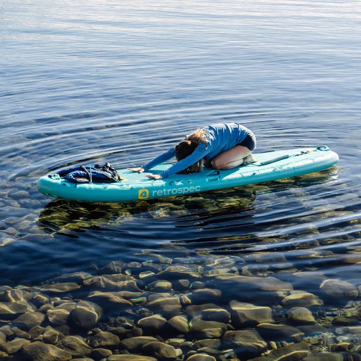 A person performs child's pose on a teal Weekender Yogi Inflatable paddleboard in calm water, with clear visibility of stones below the surface.
