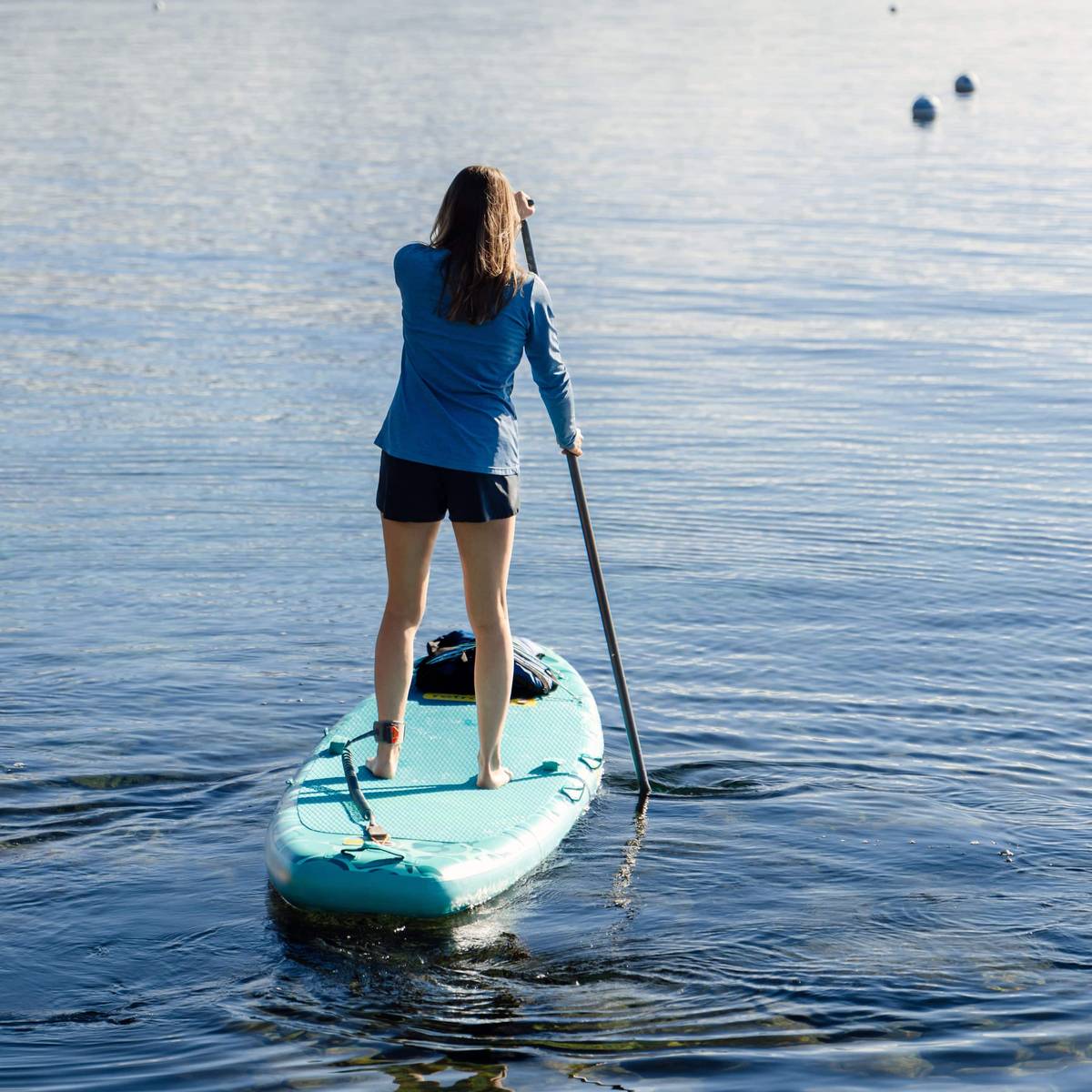 A woman paddles on the turquoise Weekender Yogi Inflatable Stand Up Paddle board on calm water under a clear sky.