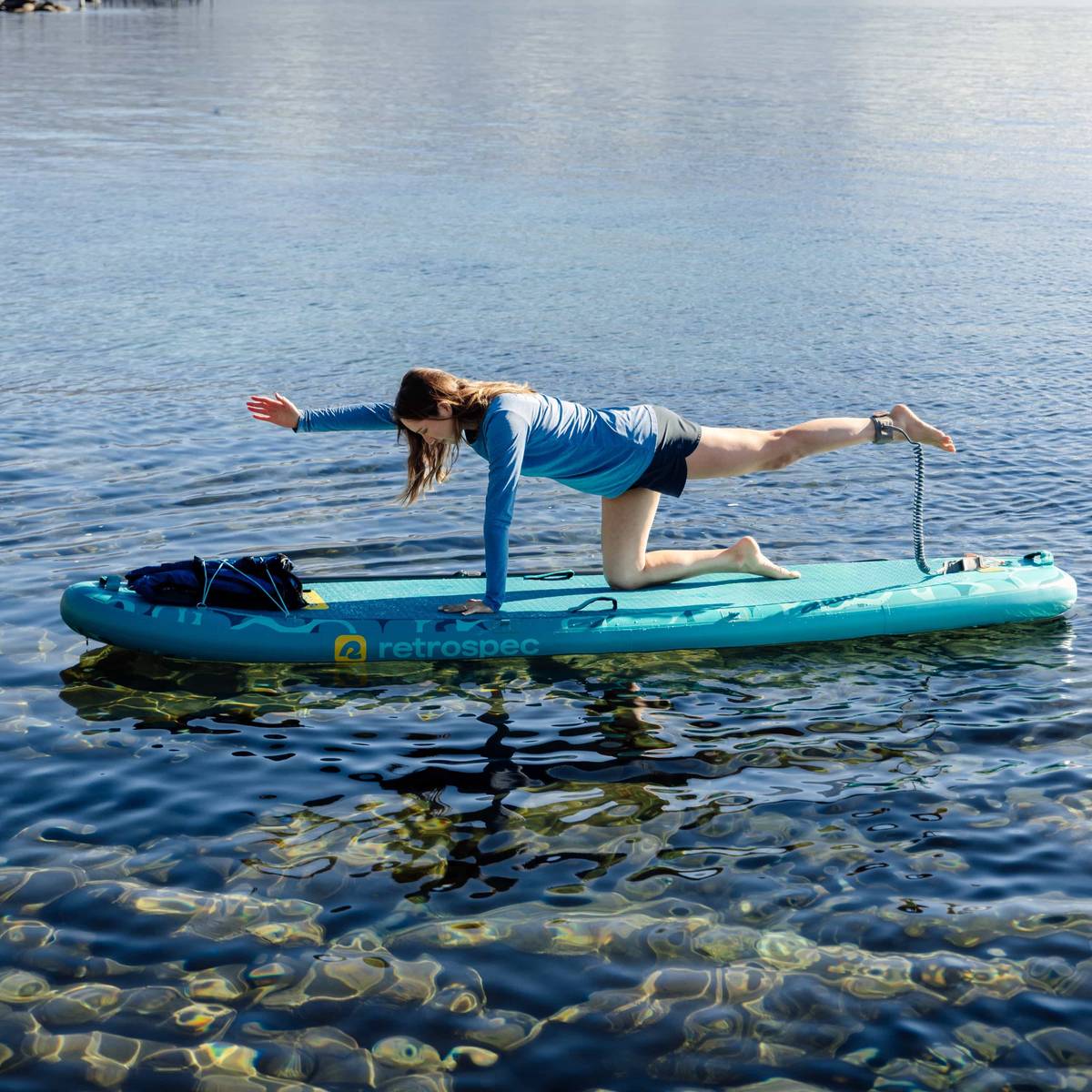 A person in a blue long-sleeve shirt performs a bird dog pose on a yoga paddleboard over clear water, surrounded by scenic nature.