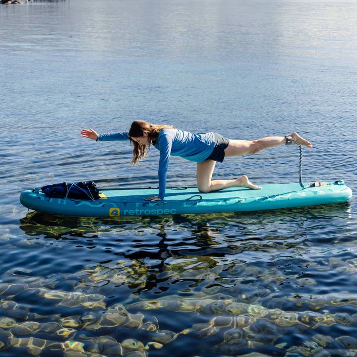 A person in a blue long-sleeve shirt performs a bird dog pose on a yoga paddleboard over clear water, surrounded by scenic nature.