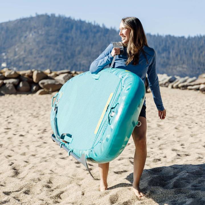 Person carrying a turquoise Weekender Yogi Inflatable Stand Up Paddle Board across a sandy beach, with mountains in the background and clear blue skies.