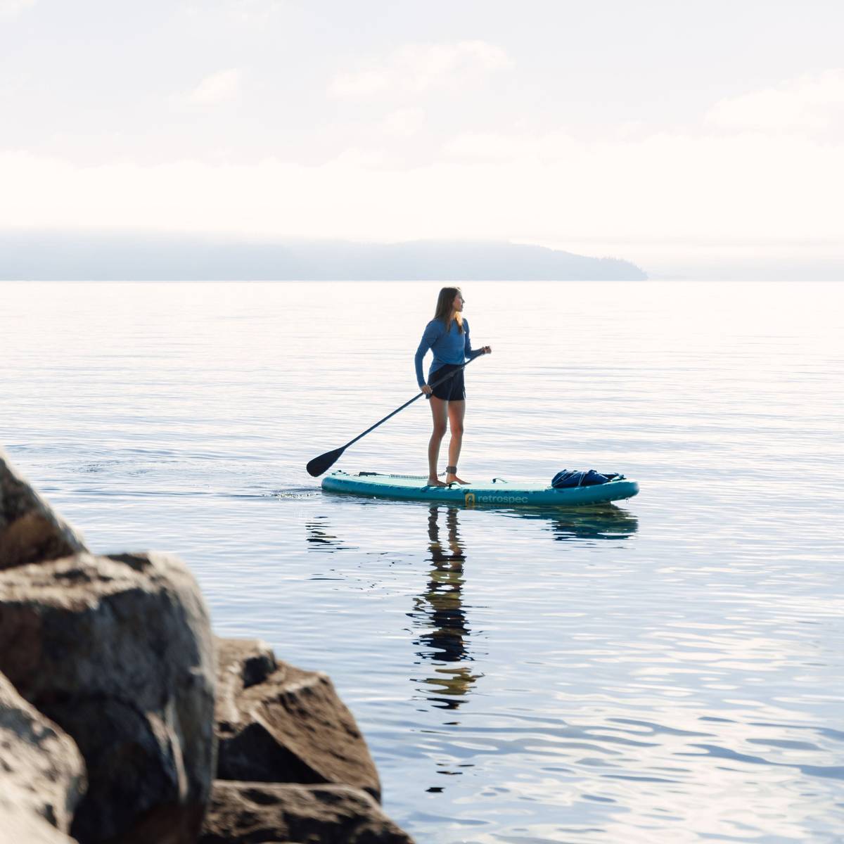 A person stands on a Inflatable Stand Up Paddle Board for yoga in calm waters, with rocks in the foreground and misty hills in the background.