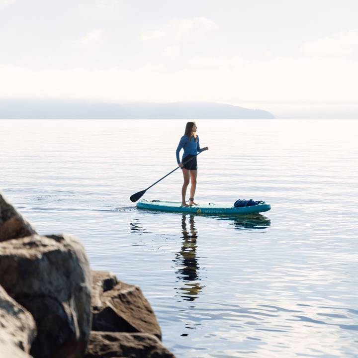 A person stands on a Inflatable Stand Up Paddle Board for yoga in calm waters, with rocks in the foreground and misty hills in the background.