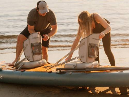 A man and woman prepare inflatable kayak seats on a paddleboard at the beach, enjoying a sunny day by the water.