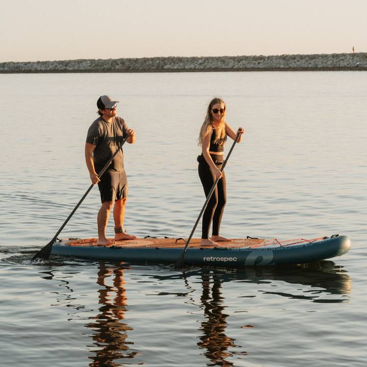 Two people paddle on calm waters on the Weekender Crew Inflatable Stand Up Paddle Board.