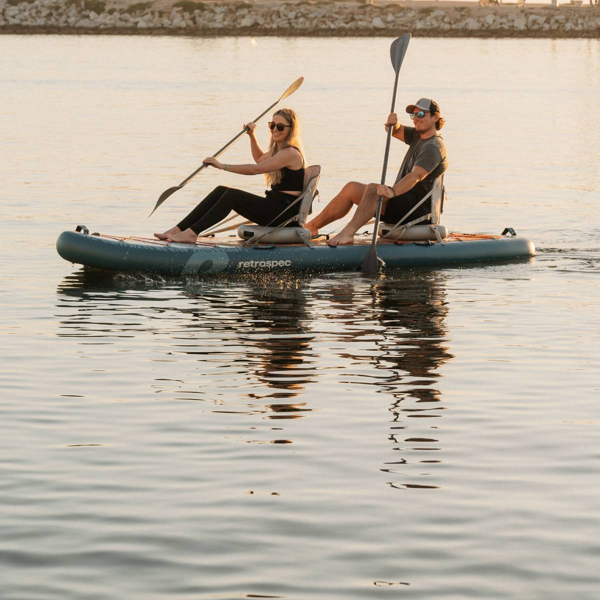 Two people paddle while sitting in inflatable kayak seats on the Weekender Crew Paddle Board in calm waters, reflecting the evening light, surrounded by a peaceful shoreline.
