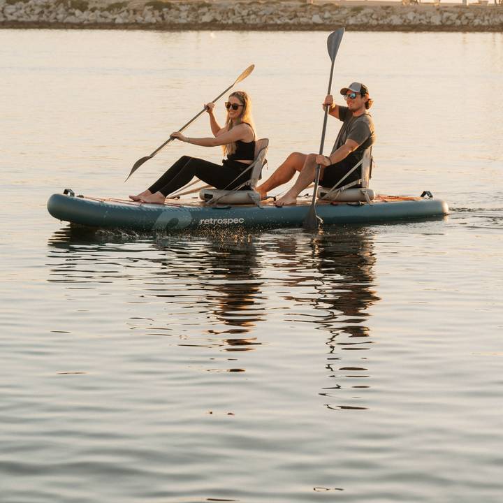 Two people paddle while sitting in inflatable kayak seats on the Weekender Crew Paddle Board in calm waters, reflecting the evening light, surrounded by a peaceful shoreline.