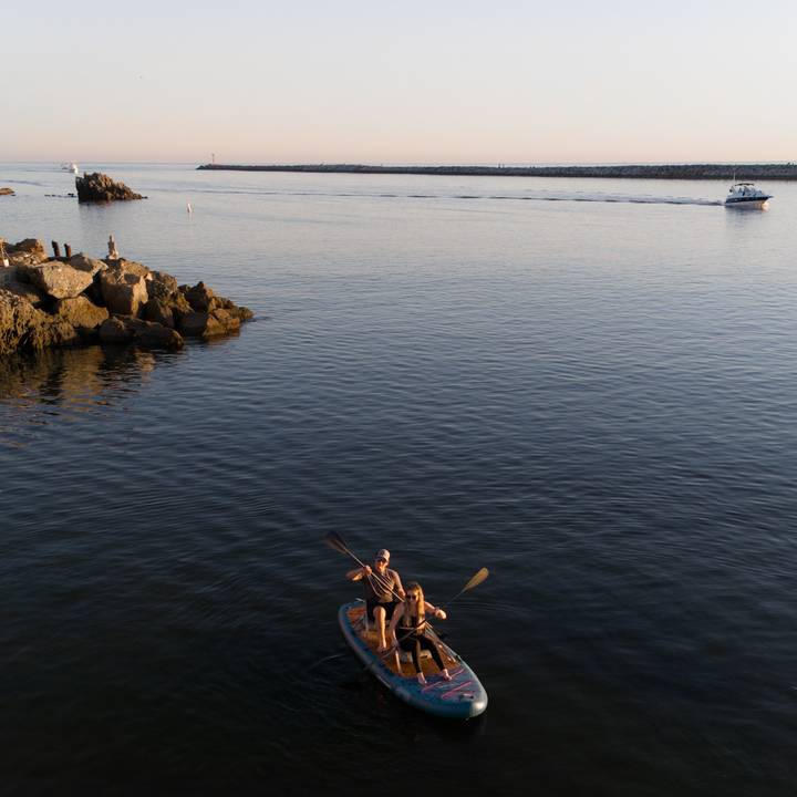 Two people site on inflatable kayak seats while paddling on the Weekender Crew Inflatable Paddle Board on calm waters with the sun setting in the distance.