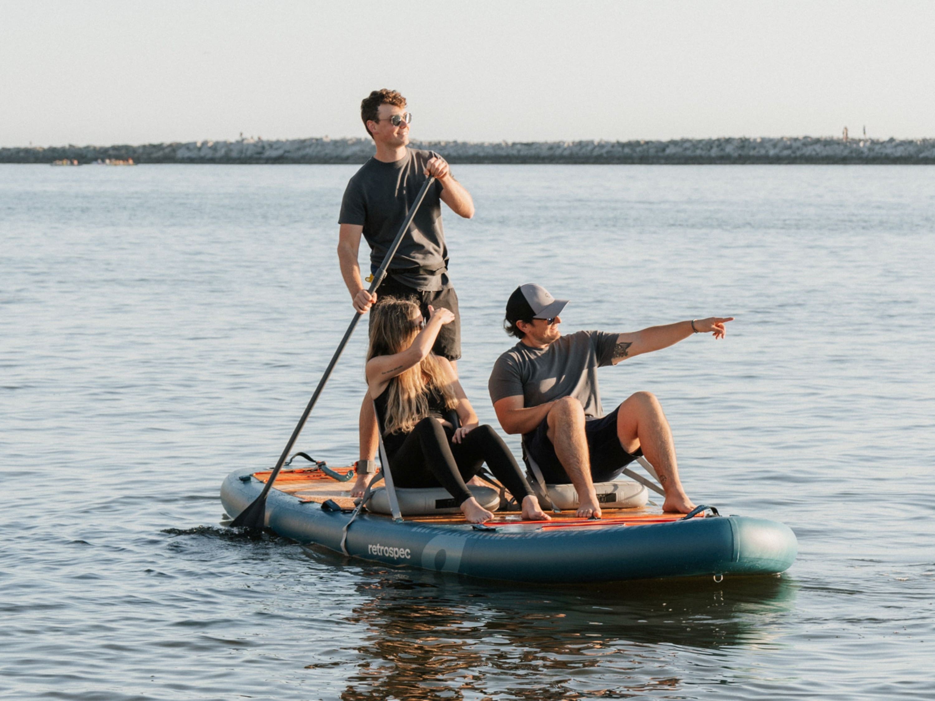 Three friends on a retrospec Weekender Crew XL inflatable Paddle Board on open water, one standing and paddling while the others sit and point toward the horizon.