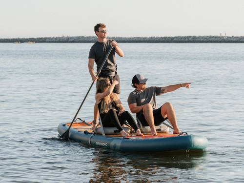 Three friends on a retrospec Weekender Crew XL inflatable Paddle Board on open water, one standing and paddling while the others sit and point toward the horizon.
