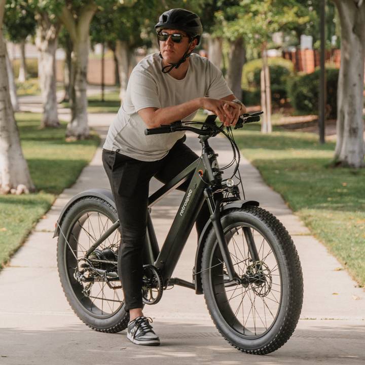 A person in a gray t-shirt and black pants leans on a matte green Koa Rev 2 Fat Tire Electric Bike, surrounded by lush greenery along a paved path.
