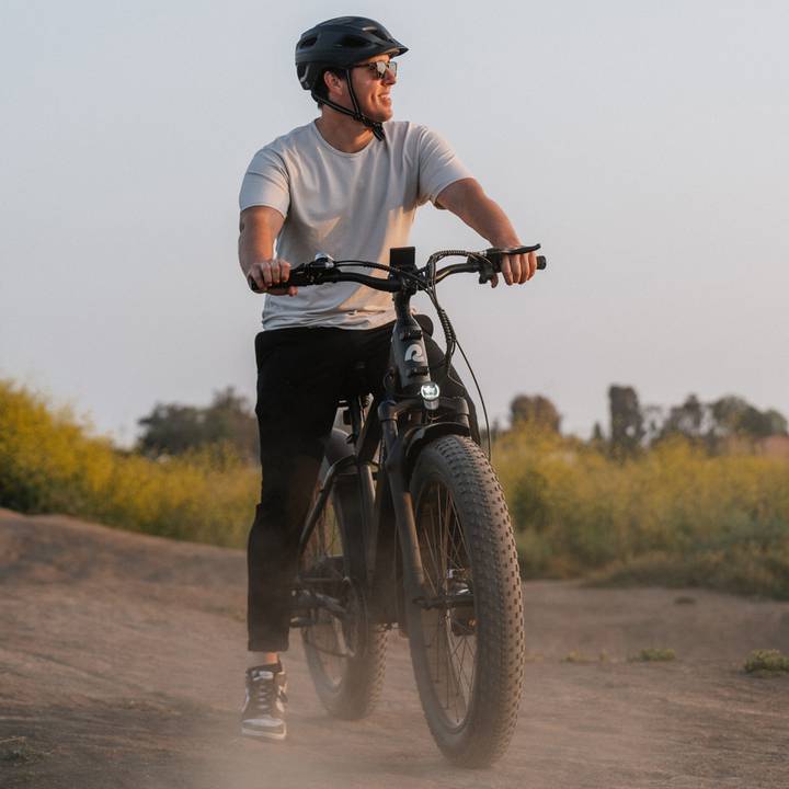 A person rides a matte green Koa Rev 2 Fat Tire Electric Bike on a dusty trail, surrounded by greenery and a soft sunset glow.