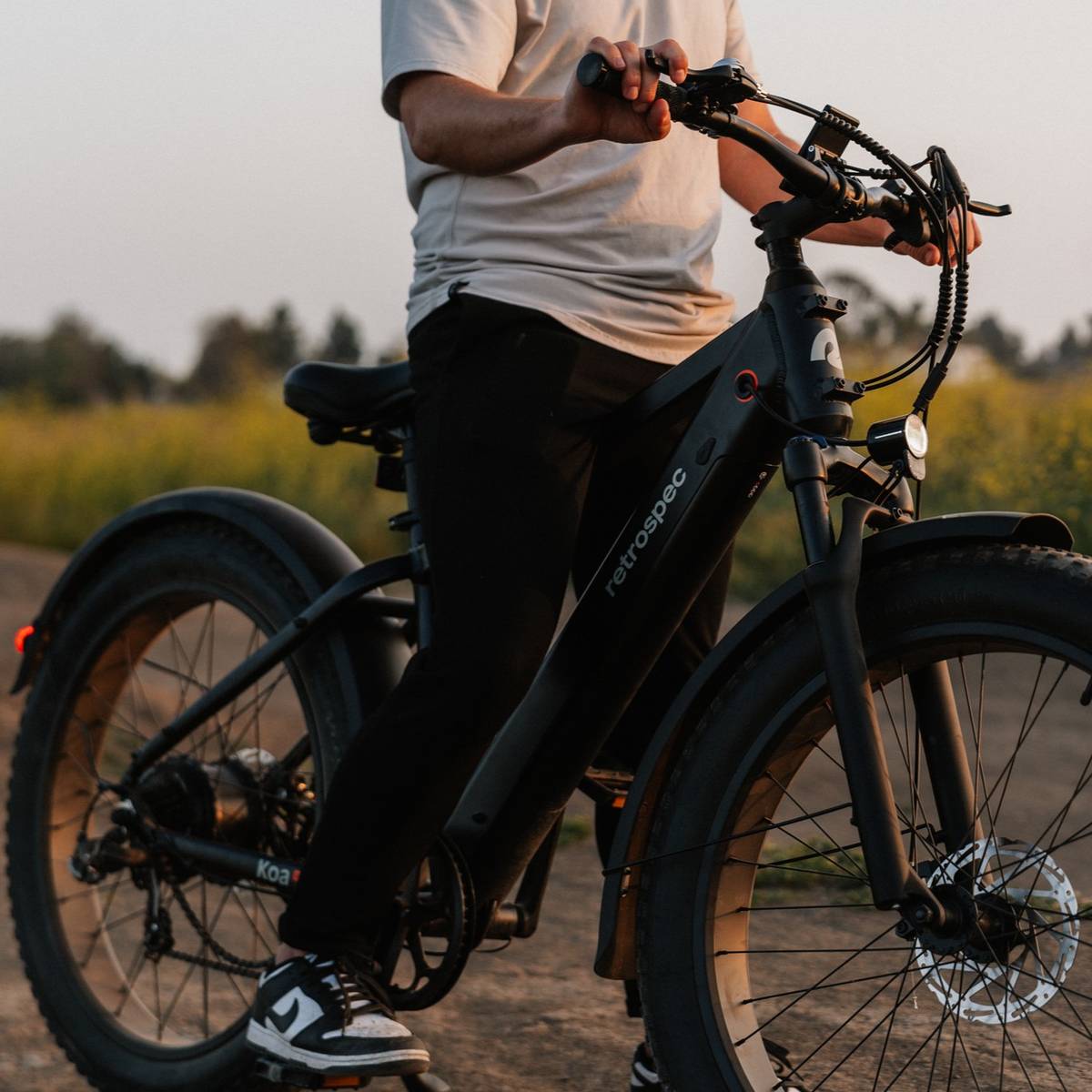 A person rides a matte black Koa Rev Fat Tire Electric Bike on a dusty trail, surrounded by greenery and a soft sunset glow.