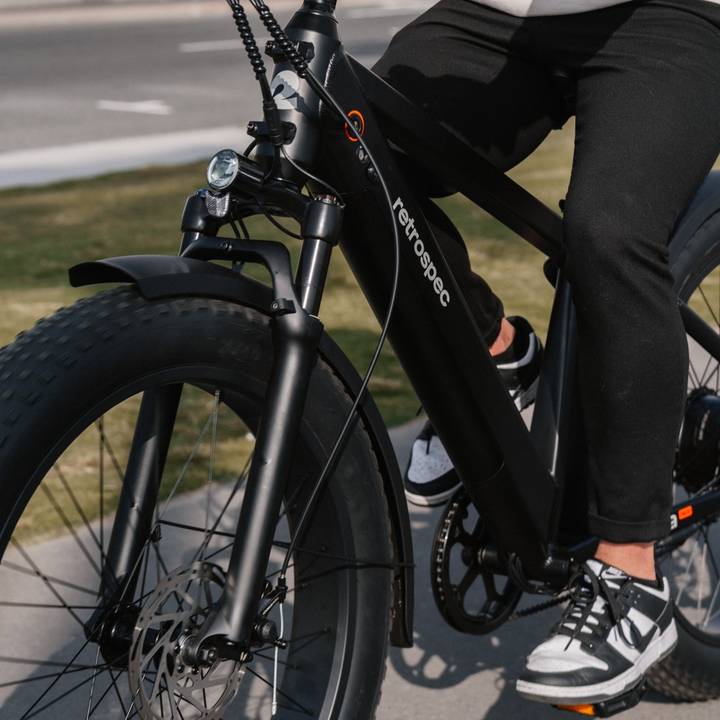 A person wearing black pants pedals a black Koa Rev Fat Tire Electric Bike, surrounded by lush greenery along a paved path.