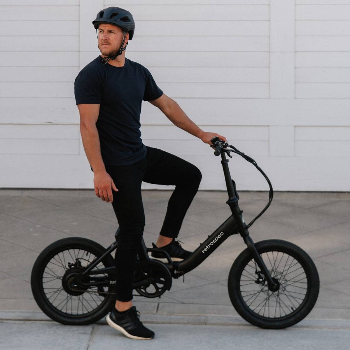 A person wearing a helmet sits on a sleek black Judd Rev Folding Electric Bike, dressed in a dark t-shirt and black pants, against a light background.