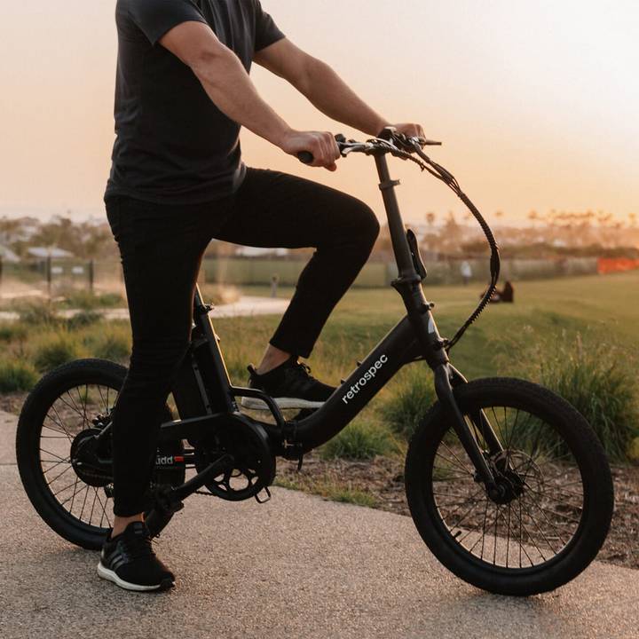 A person rides a sleek black Judd Rev Folding Electric Bike at sunset, showcasing a relaxed yet sporty urban lifestyle in a scenic park.