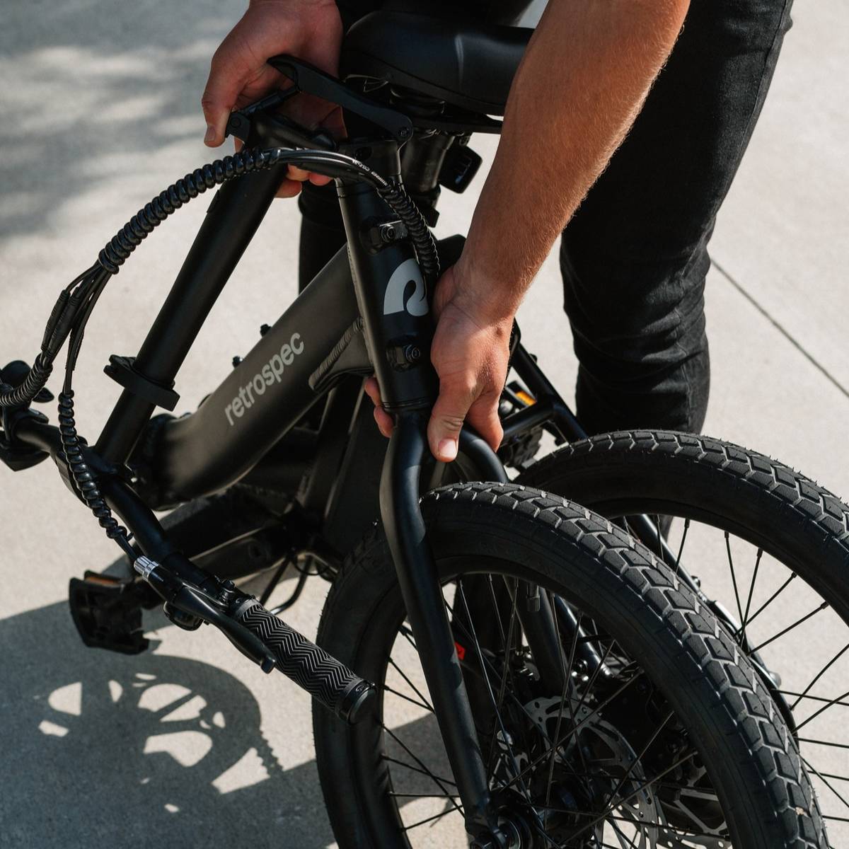 A person is holding the front fork of a folded black retrospec bicycle, preparing to lift or carry it.