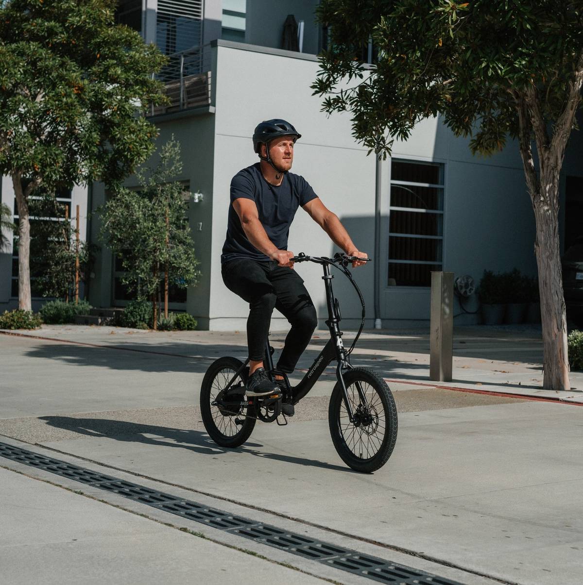 A person wearing a helmet rides a black bicycle on a sunny urban street, surrounded by trees and modern buildings.