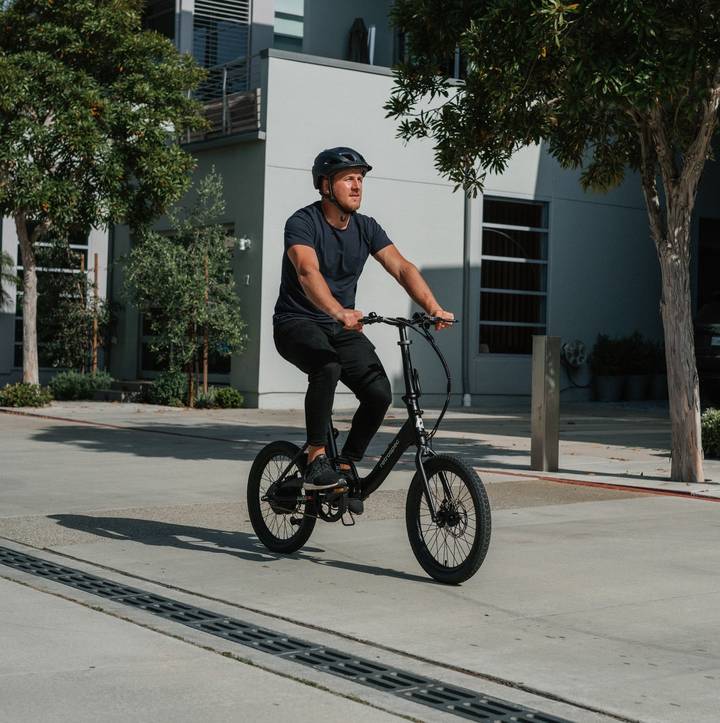 A person wearing a helmet rides a black bicycle on a sunny urban street, surrounded by trees and modern buildings.