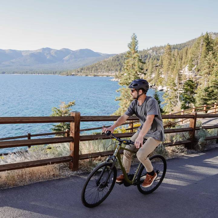 Person riding a bonsai green Barron Plus Comfort 21 Speed Hybrid Bicycle on a path near a lake with mountains and trees in the background.