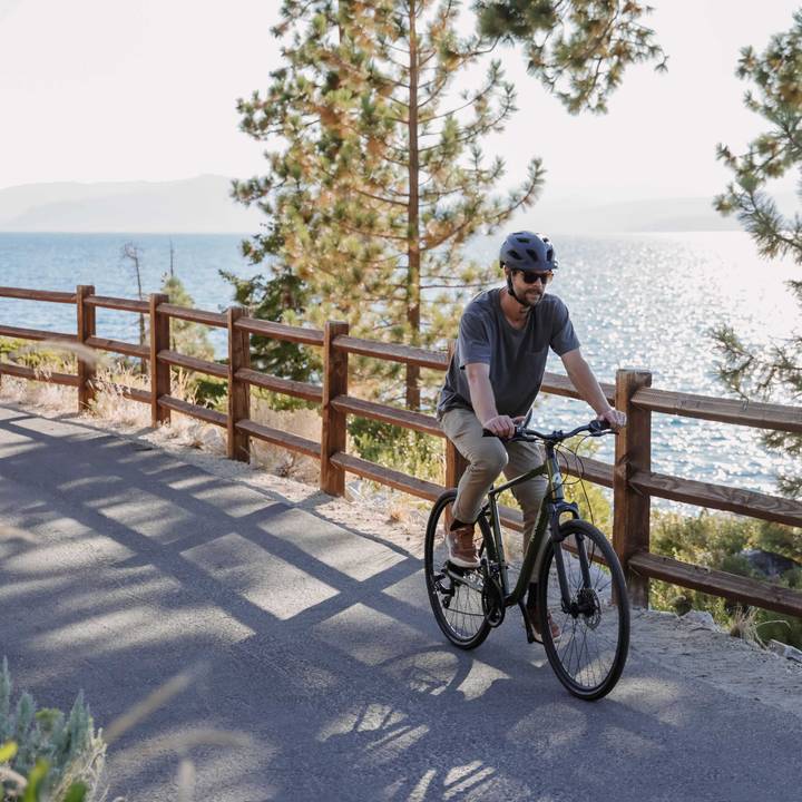 A person riding a bonsai green Barron Plus Comfort 21 Speed Hybrid Bicycle on a path beside a fence, with trees and a large body of water in the background.