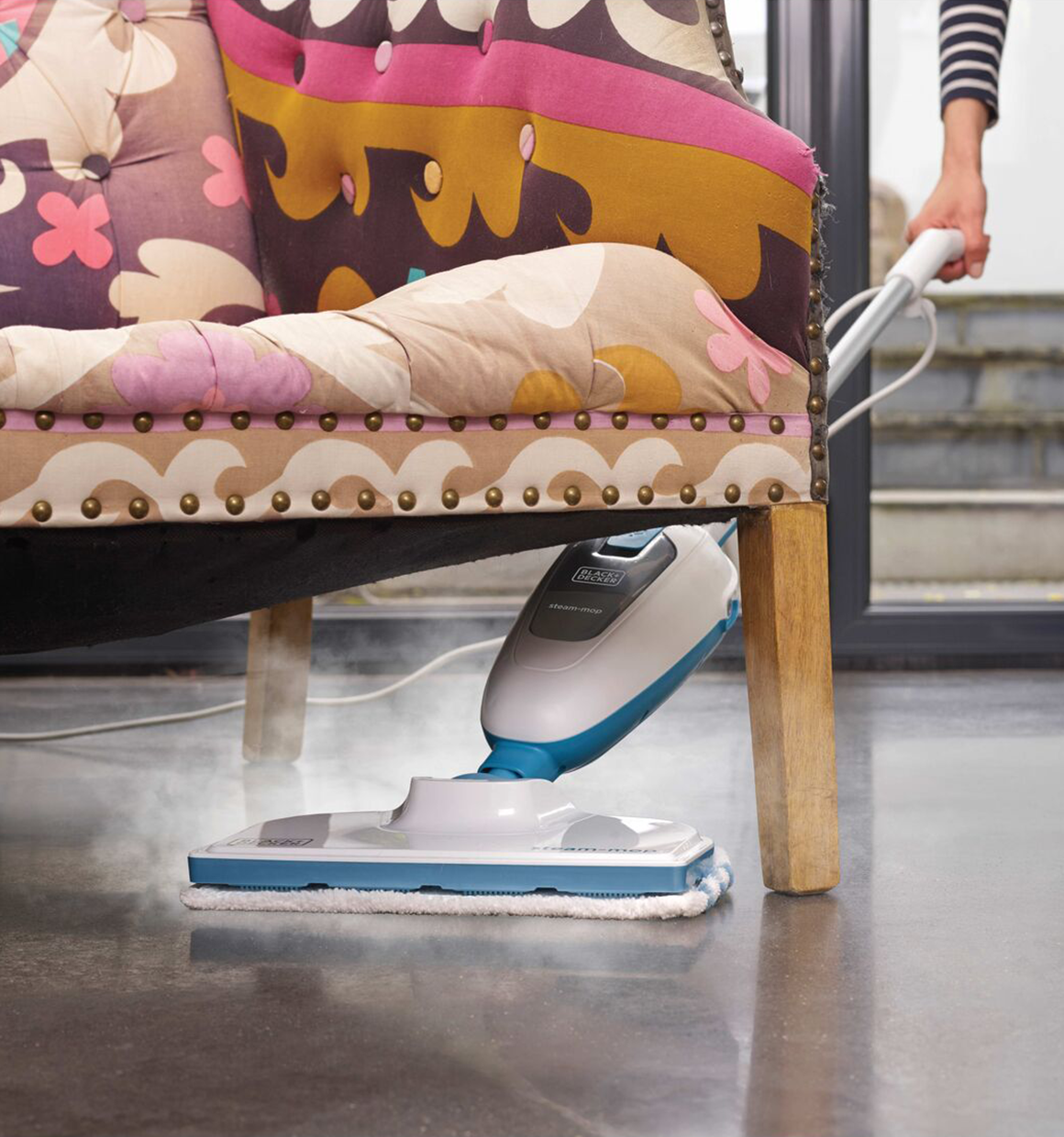 Talent using the BLACK + DECKER™ steam mop under a decorative chair, which kills up to 99% of germs