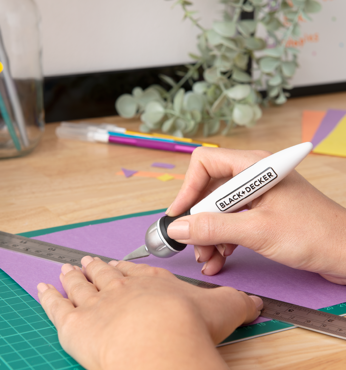 close-up of a BLACK+DECKER hobby knife being used with a rule to cut colored paper on a cutting mat