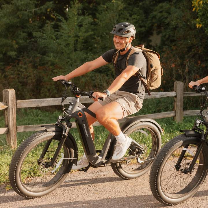 A person riding a Koa Rev+ 2 Fat Tire Electric Bike on a scenic path, wearing a helmet and a backpack, surrounded by greenery and wooden fencing.