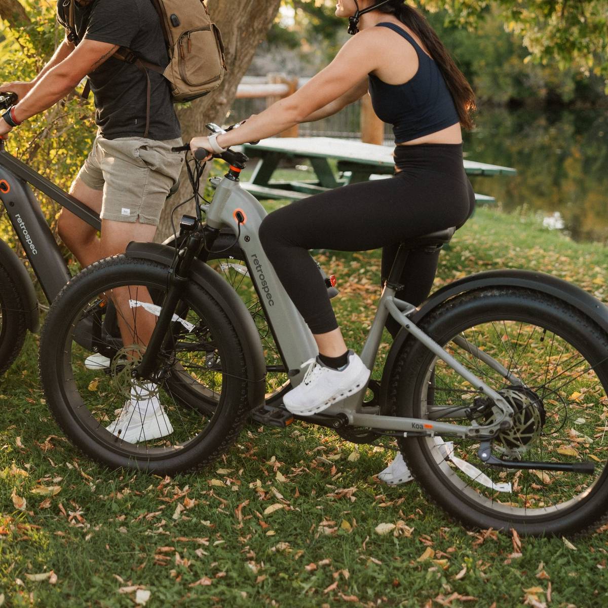 A man and woman on Koa Rev+ 2 Fat Tire Electric Bikes, dressed in casual attire, enjoy a sunny day in a park surrounded by trees and fallen leaves.
