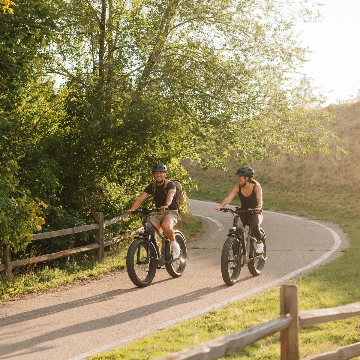 Two cyclists ride their Koa Rev+ 2 Fat Tire Electric Bikes along a winding path surrounded by greenery, enjoying a sunny day outdoors. A wooden fence borders the trail.