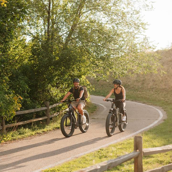 Two cyclists ride their Koa Rev+ 2 Fat Tire Electric Bikes along a winding path surrounded by greenery, enjoying a sunny day outdoors. A wooden fence borders the trail.