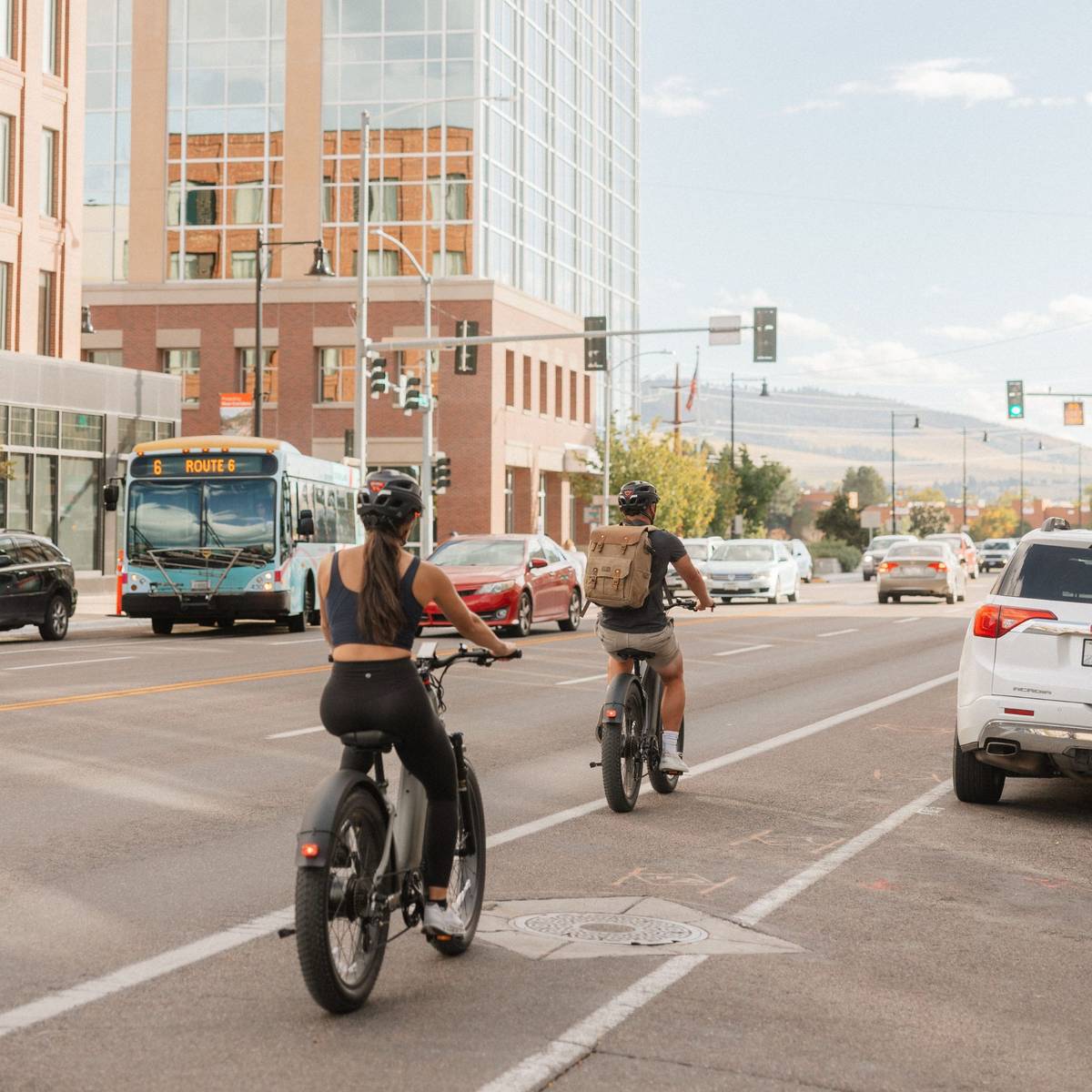 Two cyclists ride in the bike lane on their Koa Rev+ 2 Fat Tire Electric Bikes alongside cars and a bus labeled "Route 6" on a sunny urban street.