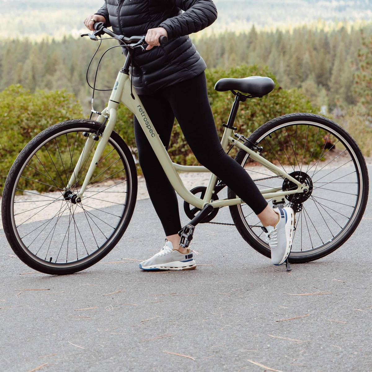 A person in a black jacket and leggings stands with a light green bicycle on a path surrounded by trees.