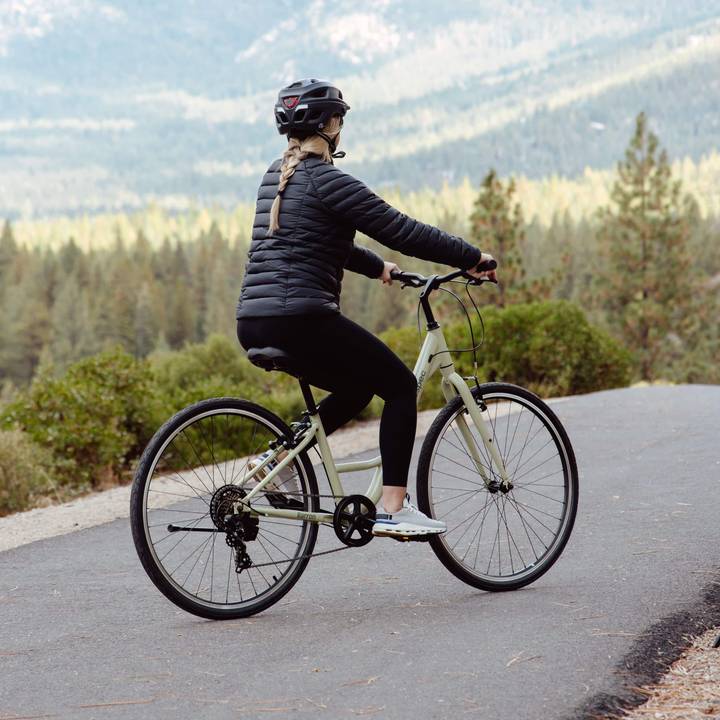 Person wearing a black helmet and jacket riding a light green bicycle on a path through a forested area.