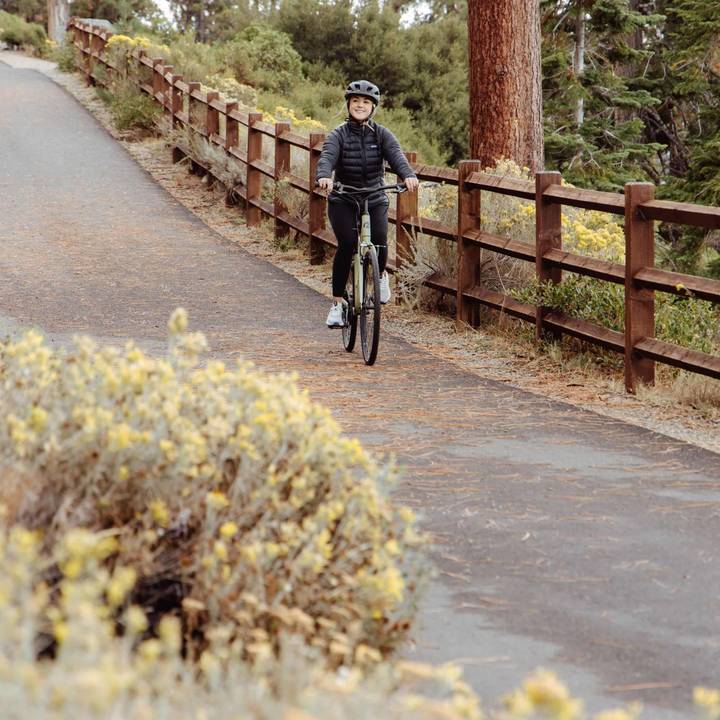 Person cycling on a paved path with greenery and wooden fence.