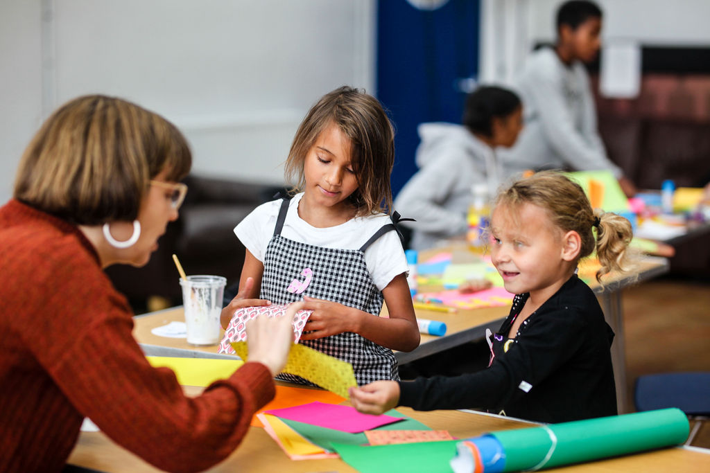 An image of a woman talking to two children who have table with coloured paper and glue between them 