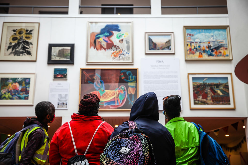 An image of a wall of artworks with the backs of four children underneath them looking up at them 