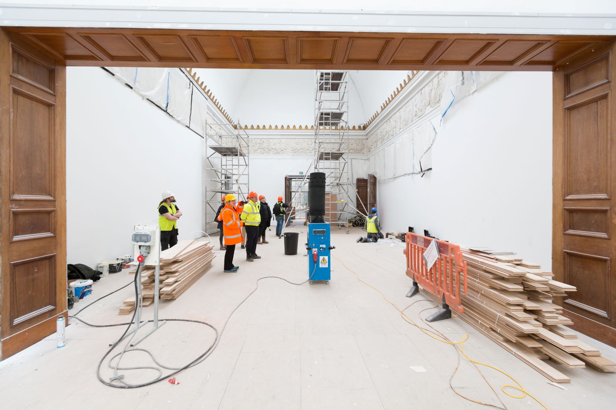 A large open gallery space, people in high vis jackets and safety helmets gather in the centre, there are wooden floorboards to the side of the room 